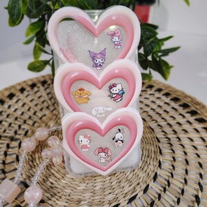 May include: A clear phone case with three heart-shaped layers, each outlined in pink and white. The case features various cartoon character stickers, including Hello Kitty and My Melody. The case is displayed on a woven mat with a pink beaded necklace.