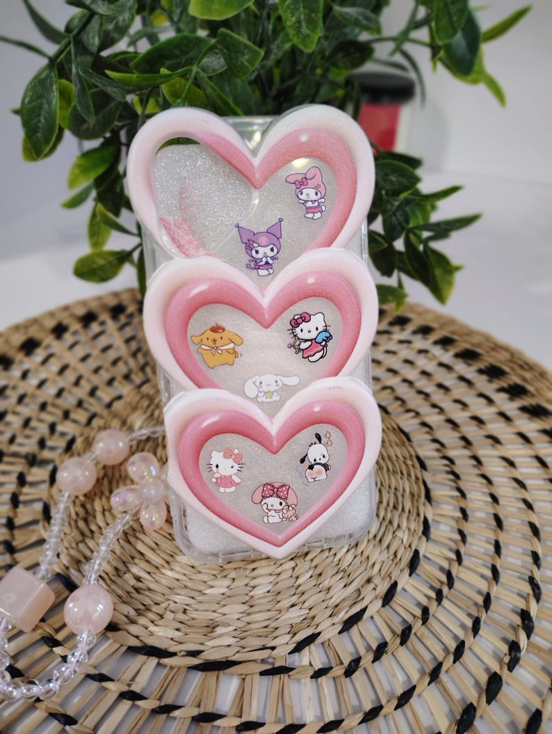 May include: A clear phone case with three heart-shaped layers, each outlined in pink and white. The case features various cartoon character stickers, including Hello Kitty and My Melody. The case is displayed on a woven mat with a pink beaded necklace.