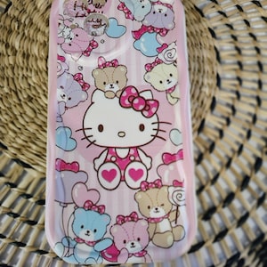 May include: Pink phone case featuring Hello Kitty and teddy bear graphics. The case has a glossy finish and is decorated with bows, hearts, and clouds. The design includes light blue, pink, and brown colors.