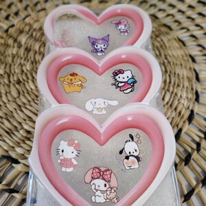 May include: A clear phone case with a heart-shaped design in pink and white. The case features multiple cartoon character stickers, including Hello Kitty, and other characters. The case is designed to hold a phone and protect it from damage.