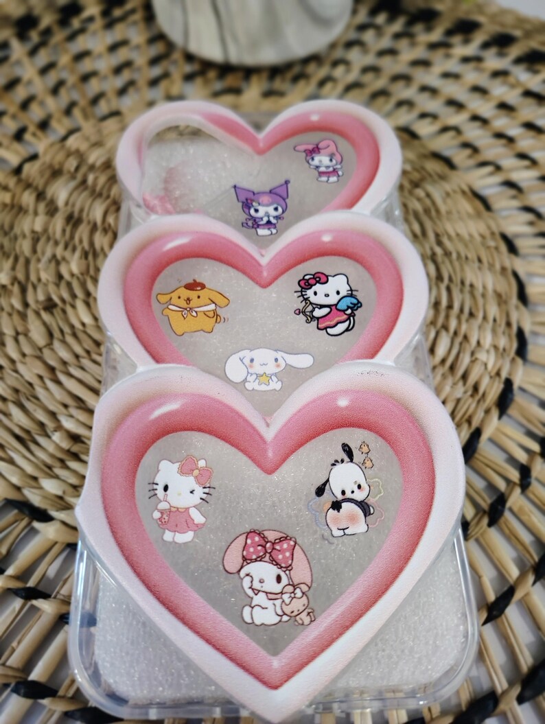May include: A clear phone case with a heart-shaped design in pink and white. The case features multiple cartoon character stickers, including Hello Kitty, and other characters. The case is designed to hold a phone and protect it from damage.