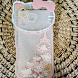 May include: Pink and clear phone case featuring a Hello Kitty design. The case has a pink bow at the top and a clear section with Hello Kitty characters and glitter. The case is shaped like Hello Kitty's head.