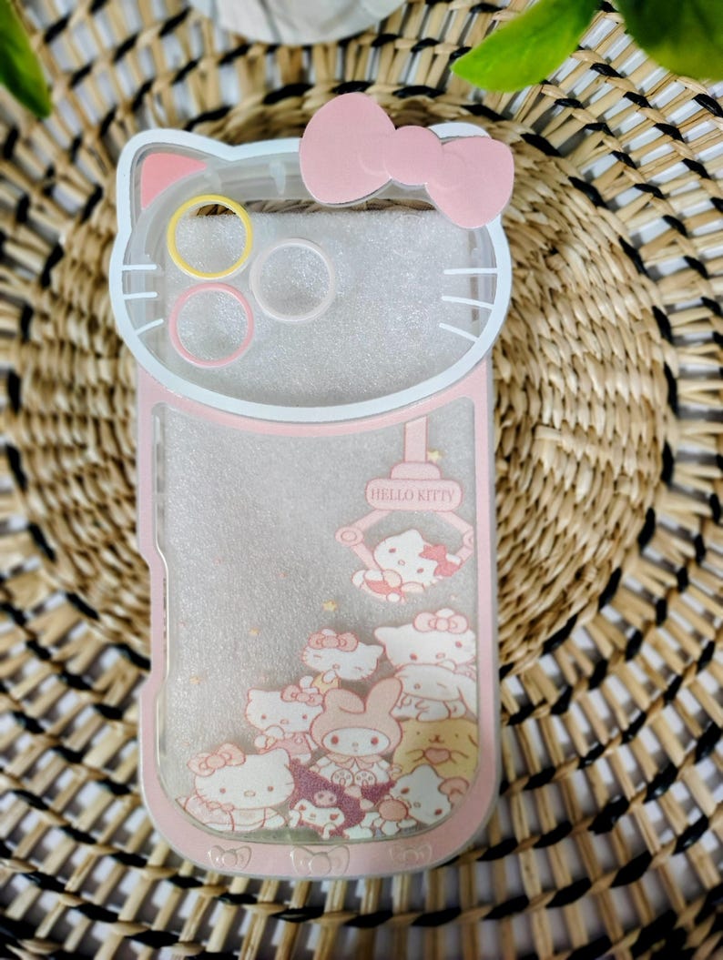 May include: Pink and clear phone case featuring a Hello Kitty design. The case has a pink bow at the top and a clear section with Hello Kitty characters and glitter. The case is shaped like Hello Kitty's head.