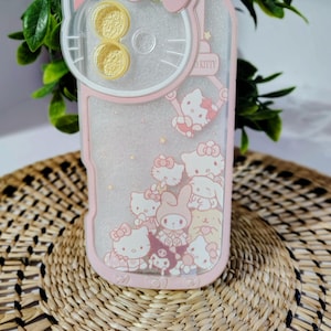 May include: Pink and clear phone case featuring Hello Kitty characters. The case has a pink border and a clear center with glitter and character designs. The top has a Hello Kitty head design with a bow and yellow accents. The case is decorated with multiple Hello Kitty characters.