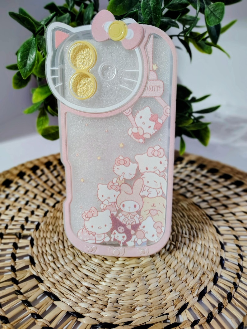 May include: Pink and clear phone case featuring Hello Kitty characters. The case has a pink border and a clear center with glitter and character designs. The top has a Hello Kitty head design with a bow and yellow accents. The case is decorated with multiple Hello Kitty characters.