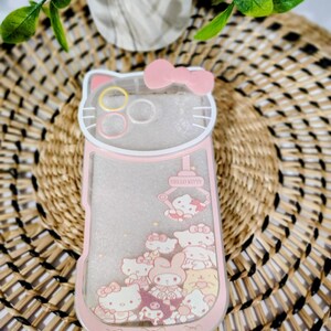 May include: A pink and clear phone case featuring a Hello Kitty design. The case has a pink bow at the top and a collection of Hello Kitty characters. The text "Hello Kitty" is visible on the case.