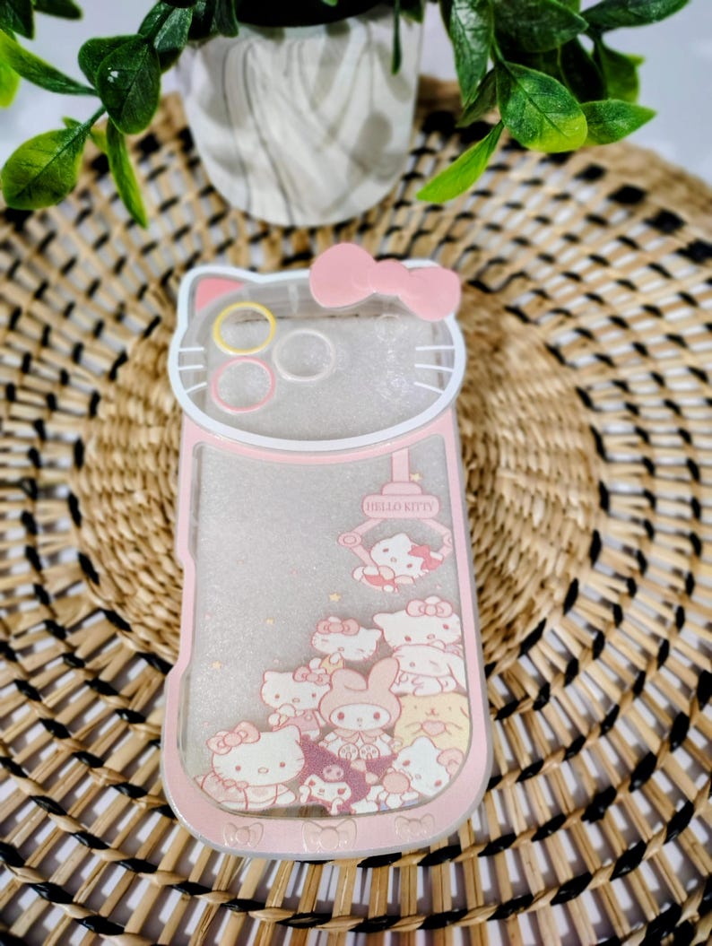 May include: A pink and clear phone case featuring a Hello Kitty design. The case has a pink bow at the top and a collection of Hello Kitty characters. The text "Hello Kitty" is visible on the case.