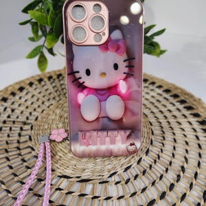 May include: A rose gold phone case with a 3D Hello Kitty design. The case has a pink strap and a small pink flower charm. The word "KITTY" is displayed at the bottom of the case.