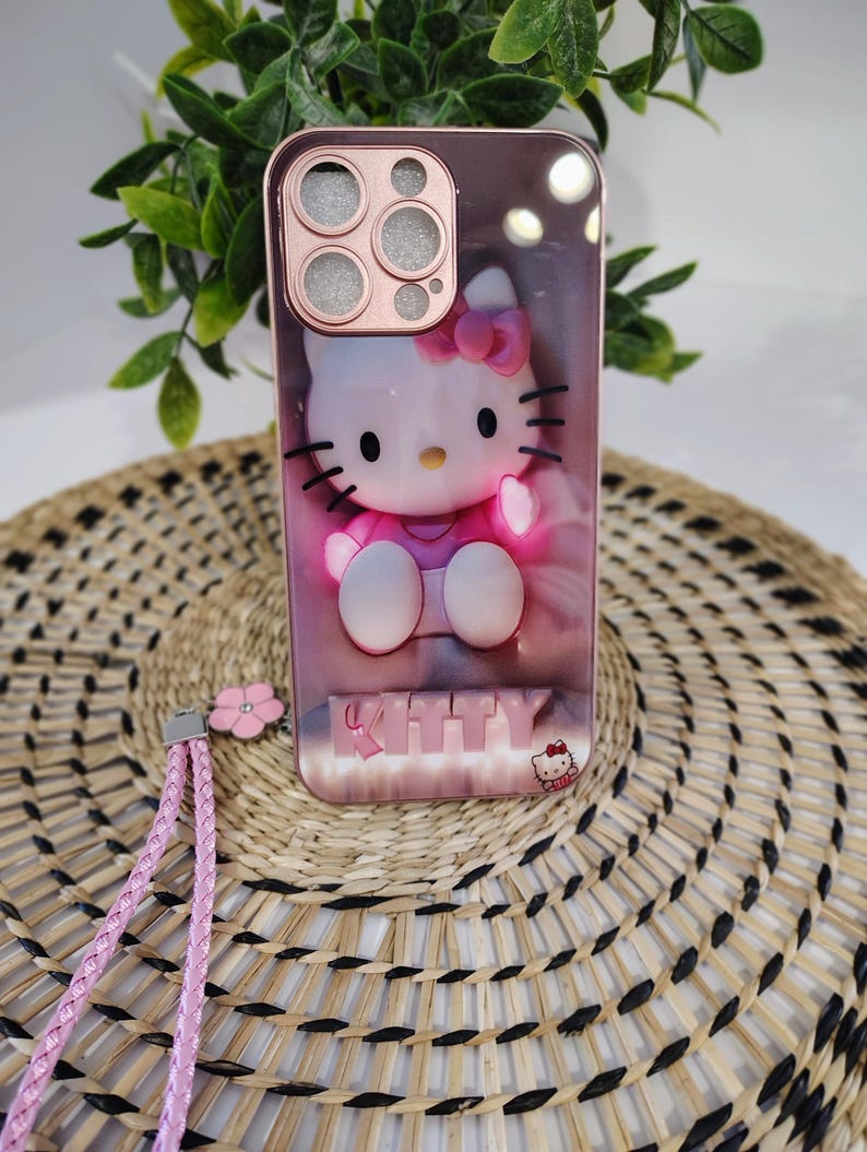 May include: A rose gold phone case with a 3D Hello Kitty design. The case has a pink strap and a small pink flower charm. The word "KITTY" is displayed at the bottom of the case.