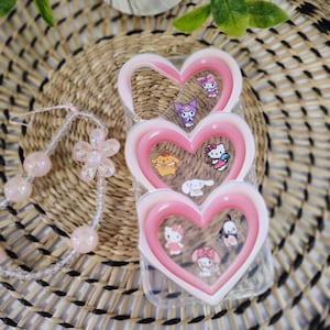 May include: A clear phone case with a pink heart-shaped frame, decorated with cartoon characters. A beaded bracelet with pink beads and a flower charm is also visible. The background includes a woven basket and greenery.