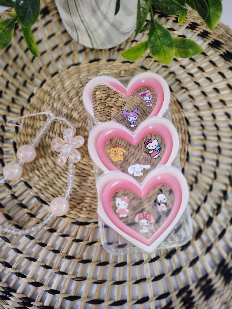 May include: A clear phone case with a pink heart-shaped frame, decorated with cartoon characters. A beaded bracelet with pink beads and a flower charm is also visible. The background includes a woven basket and greenery.