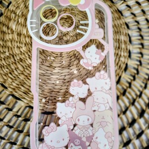May include: Pink and clear phone case featuring a Hello Kitty design. The case has a pink border and a clear center filled with Hello Kitty characters and stars. The top of the case has a Hello Kitty head shape.