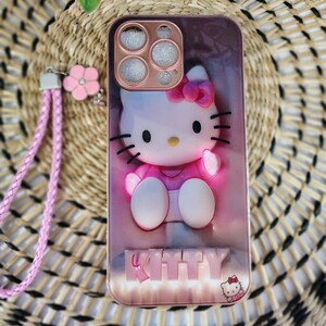 May include: A pink phone case with a 3D Hello Kitty design, complete with a pink bow and black whiskers. It includes a matching pink wrist strap and flower charm. The word "KITTY" is displayed at the bottom of the case.