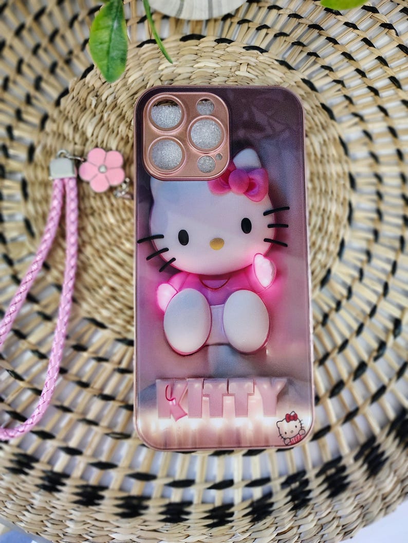 May include: A pink phone case with a 3D Hello Kitty design, complete with a pink bow and black whiskers. It includes a matching pink wrist strap and flower charm. The word "KITTY" is displayed at the bottom of the case.
