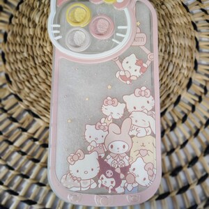 K&ouml;nnte beinhalten: Rosa-wei&szlig;es Handyh&uuml;lle mit Hello Kitty und anderen Zeichentrickfiguren. Die H&uuml;lle hat eine transparente R&uuml;ckseite, einen rosa Rand und ein Hello Kitty-Gesichtsmotiv oben. Der Text "KITTY" ist auf der Seite sichtbar.