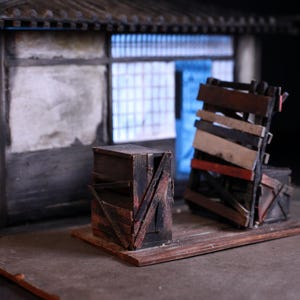 Puede incluir: Escena en miniatura con un edificio de madera envejecida, tejado de tejas y ventana azulada. Dos cajas rústicas, una vertical y otra inclinada, sobre una plataforma de madera, sugiriendo una maqueta o diorama.