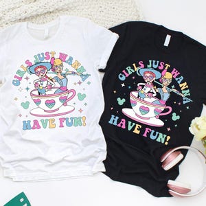 May include: Two t-shirts, one white and one black, with a vibrant graphic. The design features two cartoon characters in a teacup, accompanied by the text "GIRLS JUST WANNA HAVE FUN!" in a whimsical font. The graphic includes hearts and stars.