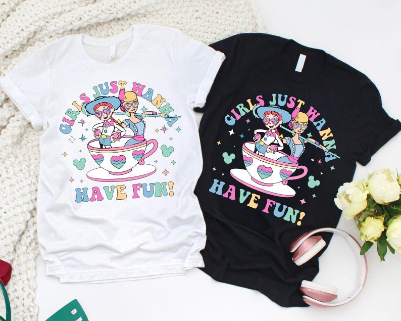 May include: Two t-shirts, one white and one black, with a vibrant graphic. The design features two cartoon characters in a teacup, accompanied by the text "GIRLS JUST WANNA HAVE FUN!" in a whimsical font. The graphic includes hearts and stars.