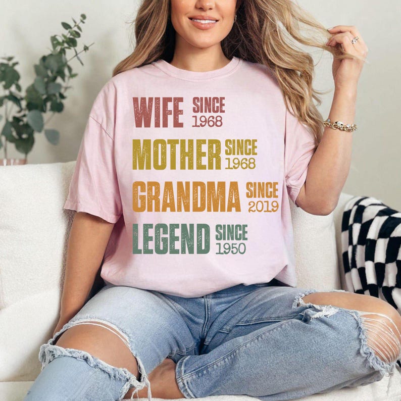 Pu&ograve; includere: Una maglietta rosa chiaro con le parole "WIFE, MOTHER, GRANDMA, LEGEND" in un design a strati. Ogni parola &egrave; di un colore diverso, con gli anni "SINCE 1968, SINCE 2019, SINCE 1950" stampati sotto. La maglietta &egrave; indossata da una persona.