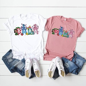 Puede incluir: Dos camisetas, una blanca y otra rosa, con la palabra "DAD" en un dise&ntilde;o de dibujos animados colorido con personajes. Las camisetas se combinan con vaqueros azules y zapatillas blancas, dispuestas sobre una superficie de madera blanca.