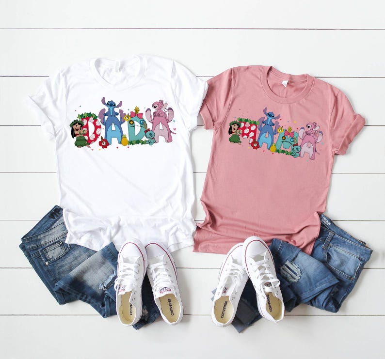 Puede incluir: Dos camisetas, una blanca y otra rosa, con la palabra "DAD" en un dise&ntilde;o de dibujos animados colorido con personajes. Las camisetas se combinan con vaqueros azules y zapatillas blancas, dispuestas sobre una superficie de madera blanca.