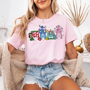 Puede incluir: Camiseta rosa claro con la palabra "Ohana" en letras de estilo c&oacute;mic. Cada letra est&aacute; dise&ntilde;ada con personajes de la pel&iacute;cula "Lilo & Stitch". La camiseta es de manga corta y cuello redondo, con un dise&ntilde;o colorido.