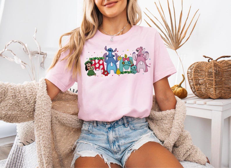 Puede incluir: Camiseta rosa claro con la palabra "Ohana" en letras de estilo c&oacute;mic. Cada letra est&aacute; dise&ntilde;ada con personajes de la pel&iacute;cula "Lilo & Stitch". La camiseta es de manga corta y cuello redondo, con un dise&ntilde;o colorido.