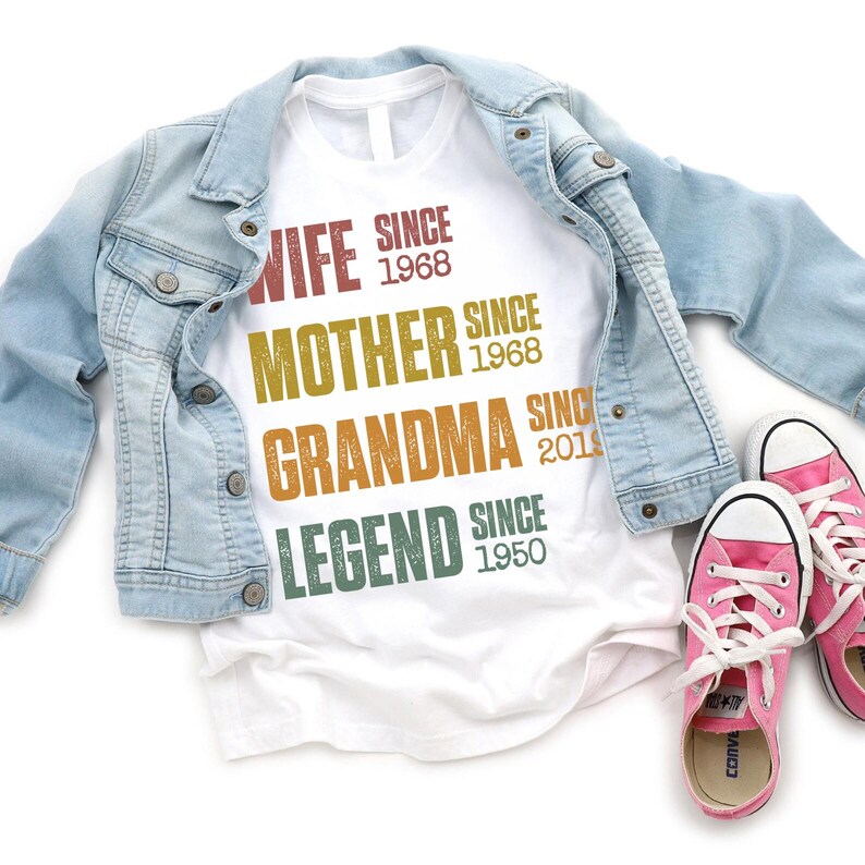 Pu&ograve; includere: Una maglietta bianca con le parole "WIFE SINCE 1968, MOTHER SINCE 1968, GRANDMA SINCE 2019, LEGEND SINCE 1950" in vari colori. La maglietta &egrave; abbinata a una giacca di jeans azzurra e scarpe da ginnastica rosa.