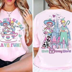 May include: Pale pink t-shirt with a vibrant graphic. The front displays two cartoon characters in a teacup, with the text "GIRLS JUST WANNA HAVE FUN!". The back features two characters in front of a castle, with the text "Walt Disney World".