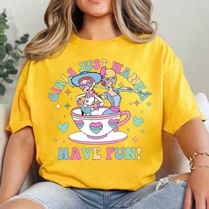 May include: Mustard yellow t-shirt with the slogan "GIRLS JUST WANNA HAVE FUN!" in a rainbow font. The design shows two cartoon figures in a teacup, with hearts and stars.