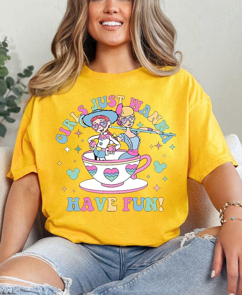 May include: Mustard yellow t-shirt with the slogan "GIRLS JUST WANNA HAVE FUN!" in a rainbow font. The design shows two cartoon figures in a teacup, with hearts and stars.