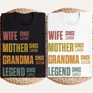Pu&ograve; includere: Due magliette, una nera e una bianca, con le parole "WIFE SINCE 1968, MOTHER SINCE 1968, GRANDMA SINCE 2019, LEGEND SINCE 1950" stampate in vari colori. Le magliette sono esposte su un cestino intrecciato.