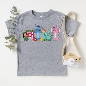 Puede incluir: Una camiseta gris de manga corta con personajes de dibujos animados coloridos que deletrean un nombre. El dise&ntilde;o incluye un personaje con falda de hierba, un extraterrestre azul y un extraterrestre rosa. Una peque&ntilde;a c&aacute;mara de madera cuelga a un lado.