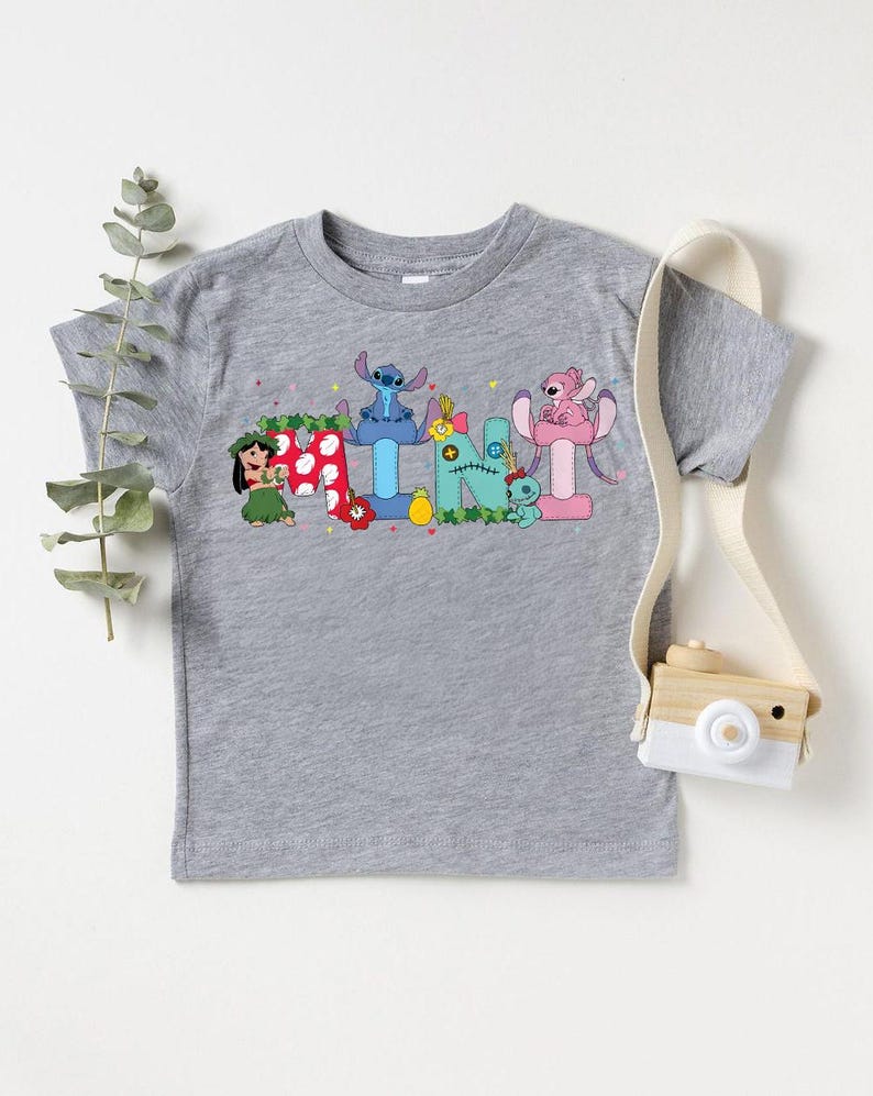 Puede incluir: Una camiseta gris de manga corta con personajes de dibujos animados coloridos que deletrean un nombre. El dise&ntilde;o incluye un personaje con falda de hierba, un extraterrestre azul y un extraterrestre rosa. Una peque&ntilde;a c&aacute;mara de madera cuelga a un lado.