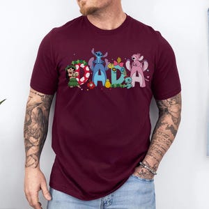 Puede incluir: Una camiseta granate con la palabra "DADA" en letras coloridas de estilo caricatura. Cada letra est&aacute; decorada con personajes de la pel&iacute;cula de animaci&oacute;n Lilo & Stitch. La camiseta es de manga corta y tiene cuello redondo.
