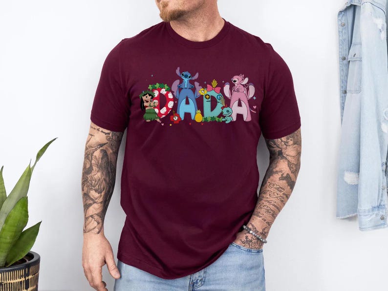 Puede incluir: Una camiseta granate con la palabra "DADA" en letras coloridas de estilo caricatura. Cada letra est&aacute; decorada con personajes de la pel&iacute;cula de animaci&oacute;n Lilo & Stitch. La camiseta es de manga corta y tiene cuello redondo.