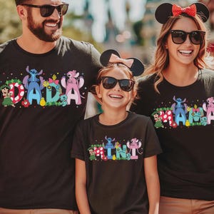 Puede incluir: Tres camisetas negras con gr&aacute;ficos coloridos. Las camisetas presentan las palabras "DADA", "MAMA" y "MINI" con personajes de dibujos animados. Las camisetas son usadas por una familia, cada uno con gafas de sol y orejas de rat&oacute;n.