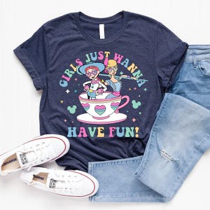 May include: Navy blue t-shirt featuring a vibrant graphic of two cartoon characters in a teacup, with the text "GIRLS JUST WANNA HAVE FUN!". The shirt is styled with light blue jeans and white Converse trainers.