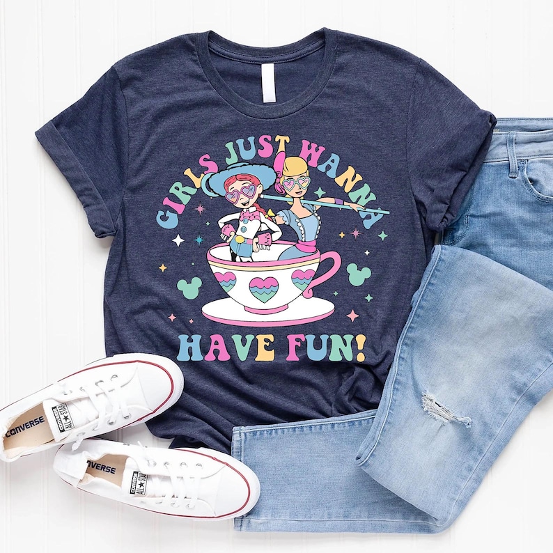 May include: Navy blue t-shirt featuring a vibrant graphic of two cartoon characters in a teacup, with the text "GIRLS JUST WANNA HAVE FUN!". The shirt is styled with light blue jeans and white Converse trainers.