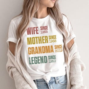Pu&ograve; includere: T-shirt bianco con le scritte "WIFE SINCE 1968", "MOTHER SINCE 1968", "GRANDMA SINCE 2019" e "LEGEND SINCE 1950" stampate in vari colori. La maglietta &egrave; indossata.
