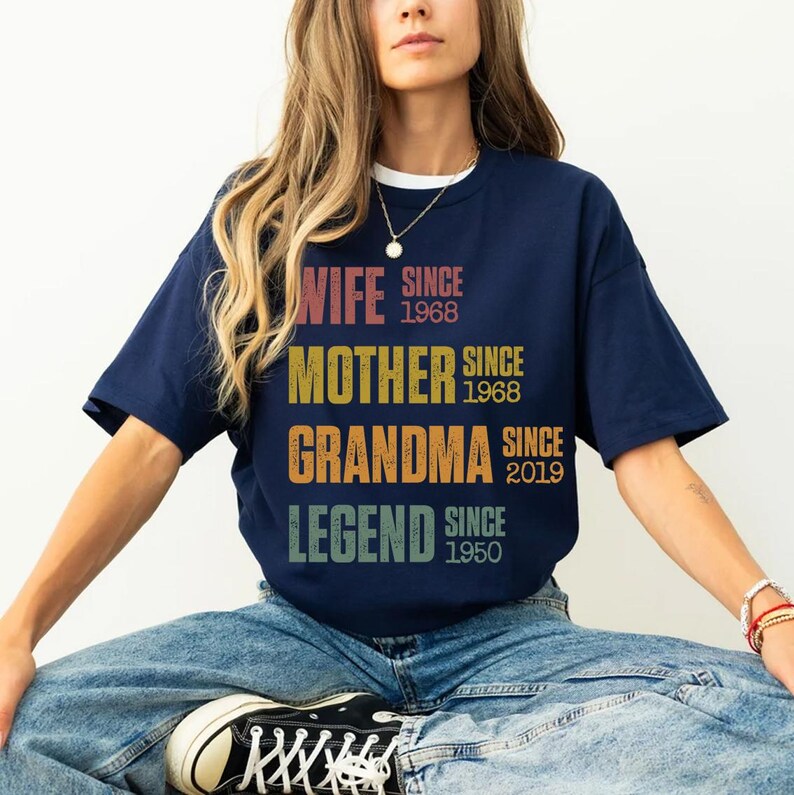 Pu&ograve; includere: T-shirt blu navy con le parole "WIFE, MOTHER, GRANDMA, LEGEND" in un font retr&ograve;. La maglietta include anche gli anni 1968, 1968, 2019 e 1950. La maglietta ha un colletto bianco.
