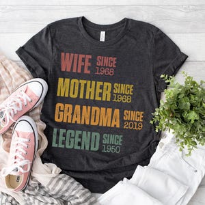 Pu&ograve; includere: T-shirt grigio scuro con le parole "WIFE SINCE 1968, MOTHER SINCE 1968, GRANDMA SINCE 2019, LEGEND SINCE 1950" in testo colorato. Scarpe da ginnastica rosa e pantaloni bianchi accanto alla maglietta. Una piccola pianta verde &egrave; anche nell'immagine.