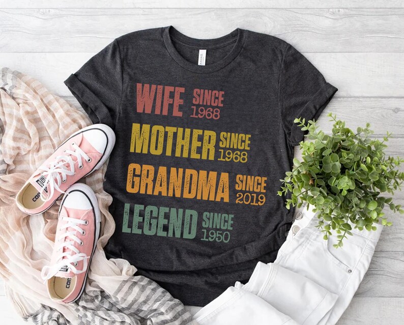Pu&ograve; includere: T-shirt grigio scuro con le parole "WIFE SINCE 1968, MOTHER SINCE 1968, GRANDMA SINCE 2019, LEGEND SINCE 1950" in testo colorato. Scarpe da ginnastica rosa e pantaloni bianchi accanto alla maglietta. Una piccola pianta verde &egrave; anche nell'immagine.