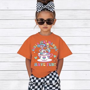 May include: An orange t-shirt with the text "GIRLS JUST WANNA HAVE FUN!" and a graphic of two girls in a teacup. The child is wearing sunglasses, a black and white checkered bow, and checkered shorts.