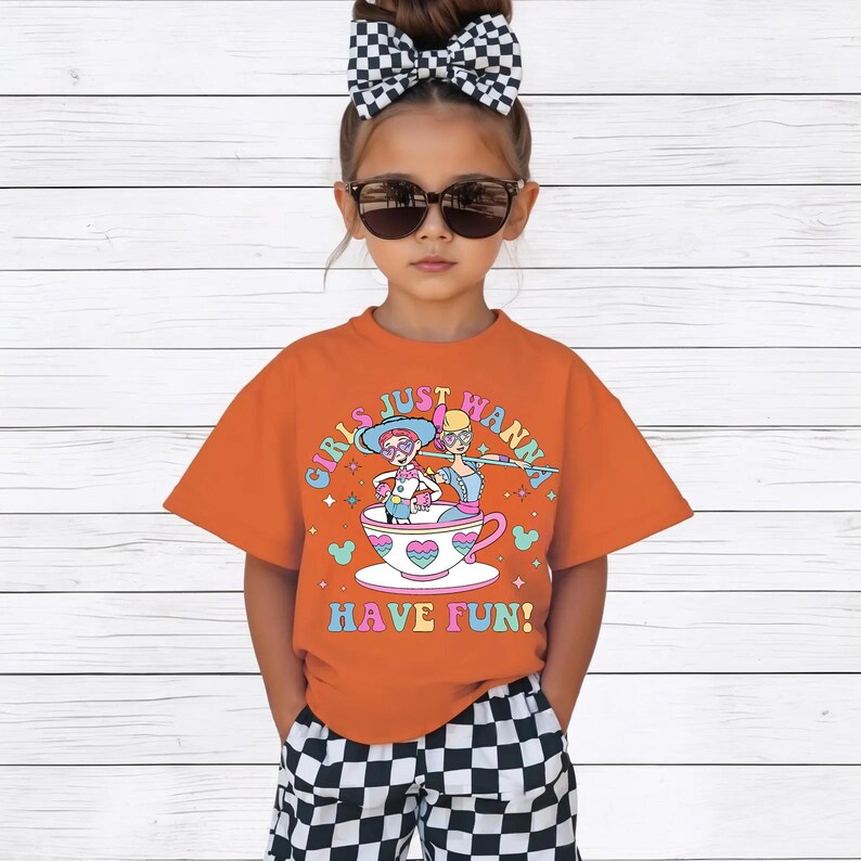 May include: An orange t-shirt with the text "GIRLS JUST WANNA HAVE FUN!" and a graphic of two girls in a teacup. The child is wearing sunglasses, a black and white checkered bow, and checkered shorts.