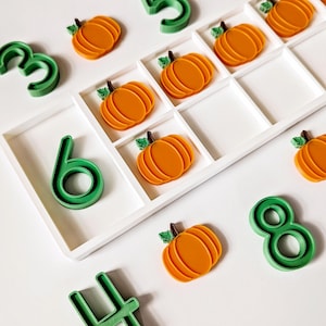 May include: A white ten-frame tray containing several orange pumpkin-shaped counters with green stems. Scattered around the tray are green plastic number molds representing the digits three, four, five, six, and eight, used for counting and math activities.