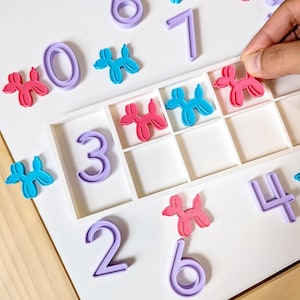 May include: A white ten-frame tray containing purple plastic numbers and small pink and blue balloon dog figures. The set includes individual numbers zero through seven and multiple balloon dog shapes arranged on a flat surface.