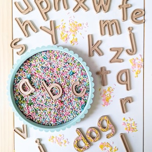 May include: A light blue scalloped tray filled with colorful sprinkles and the letters a, b, and c. Surrounding the tray are various tan plastic lowercase alphabet letters and scattered sprinkles on a white surface.