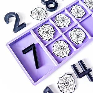 May include: A light purple rectangular tray with multiple compartments containing several white circular tokens featuring black spider web illustrations, accompanied by scattered black plastic numbers including 2, 7, 8, 0, 4, and 1.