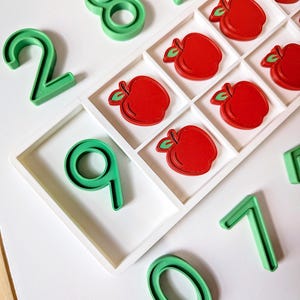 May include: A white ten-frame tray containing five red apple-shaped counters, surrounded by several green plastic number figures including 0, 1, 2, 7, 8, and 9. The set is designed for educational counting and basic mathematics practice.